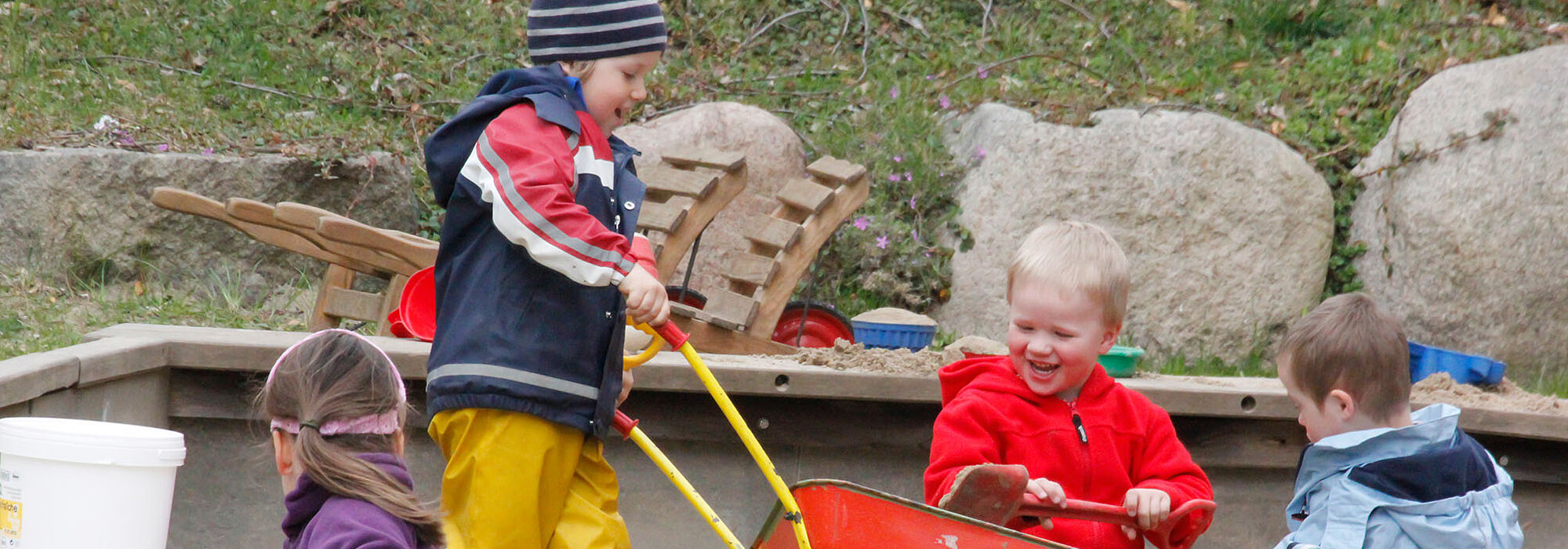Vier KInder spielen in der Sandkiste mit einer Schubkarre.